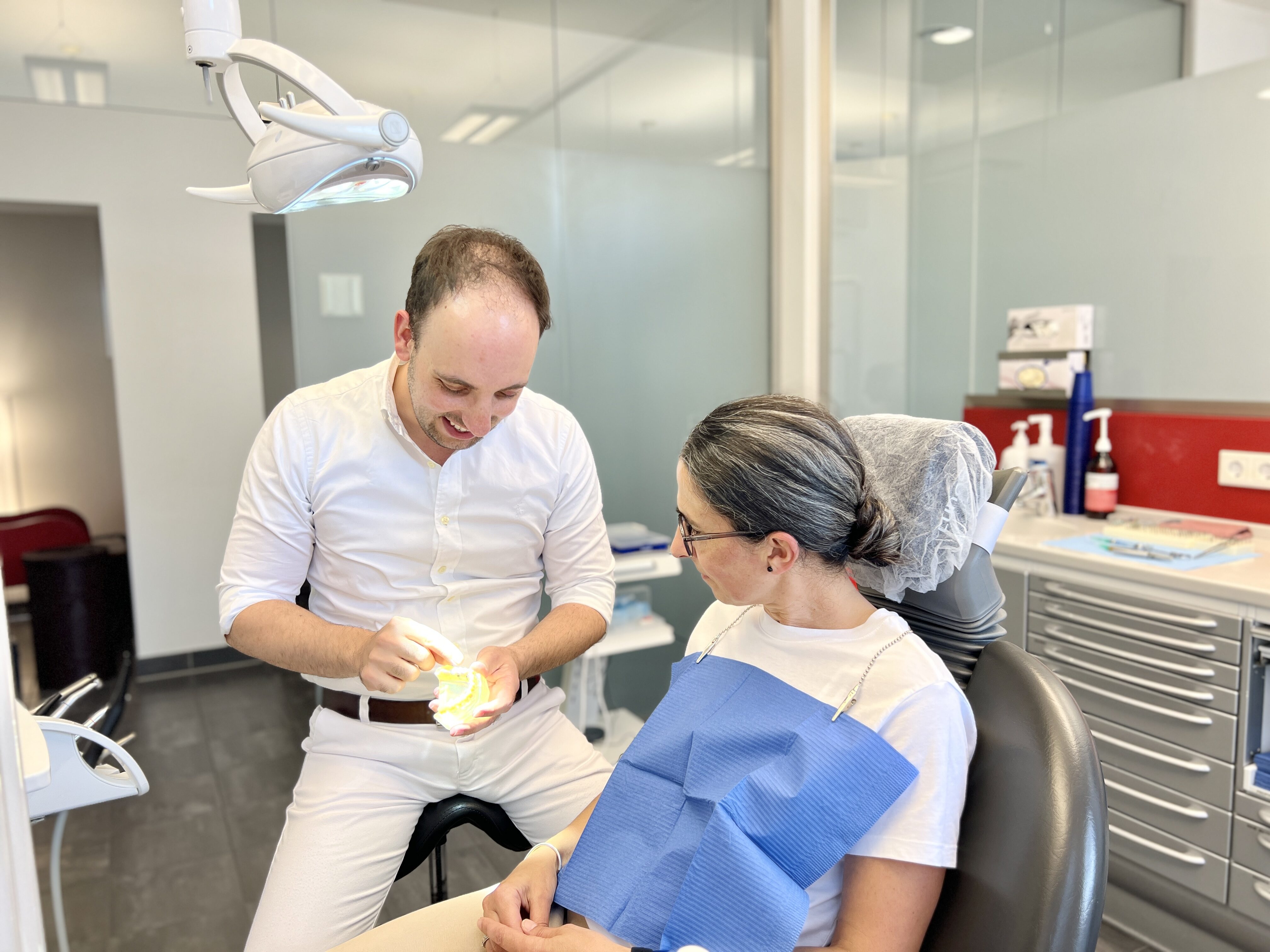 Ein Zahnarzt erklärt einer Patientin lächelnd etwas, beide in weiß gekleidet.